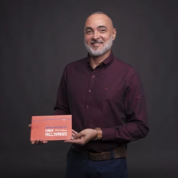 Foto de Lucio Pessoa, homem branco de barba grisalha, sorrindo. Ele veste uma camisa social vinho e segura seu livro de capa laranja intitulado 'Poemas Horizontais para Mundos Inclinados'. O fundo da imagem é cinza escuro, em um retrato profissional de estúdio.